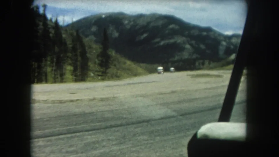 8mm home movie frame from a moving vehicle in Summit County, Colorado, 1962, showing two white cars on a wide mountain road flanked by evergreens and distant peaks.