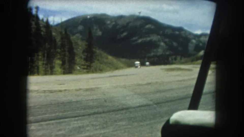 Mid-clip 8mm frame from a 1962 Summit County road trip showing the wide mountain road with forested slopes and two small white vehicles in the middle distance.