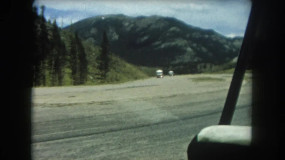 Final frame of a 1962 Summit County 8mm clip, forward-facing in-car view of a Rocky Mountain road with conifers, distant white vehicles, and a forested mountain backdrop.