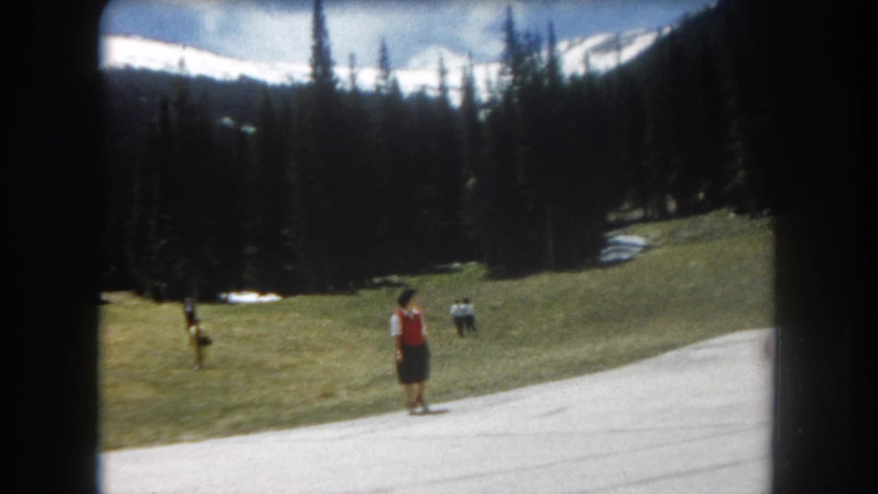 8mm home movie frame, c.1962, Summit County, Colorado — woman in red vest on paved path with snow-capped Rocky Mountain peaks and dense conifers behind.