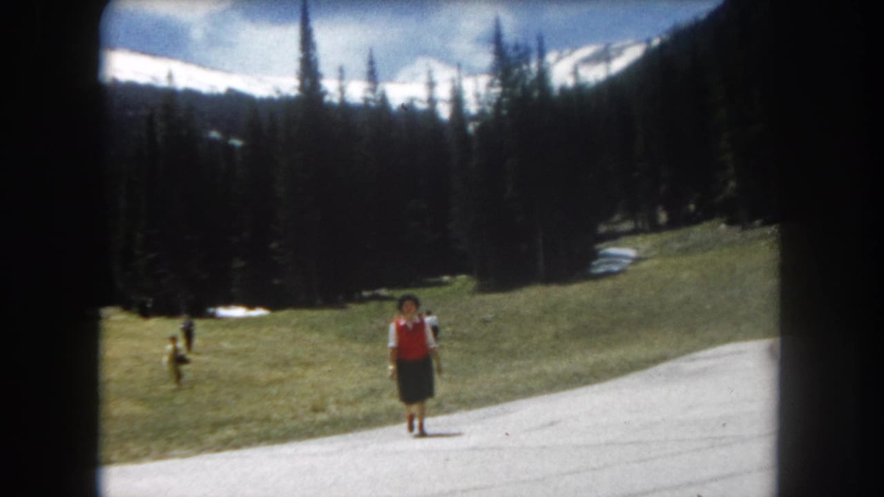 8mm home movie frame, c.1962, Summit County, Colorado — woman in red vest advancing along paved mountain path, Rocky Mountain peaks and tree line behind.