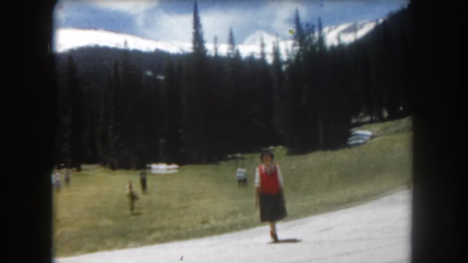 8mm home movie frame, c.1962, Summit County, Colorado — woman in red vest on path in wide mountain panorama showing snow-capped peaks, conifers, and lingering snow patches.