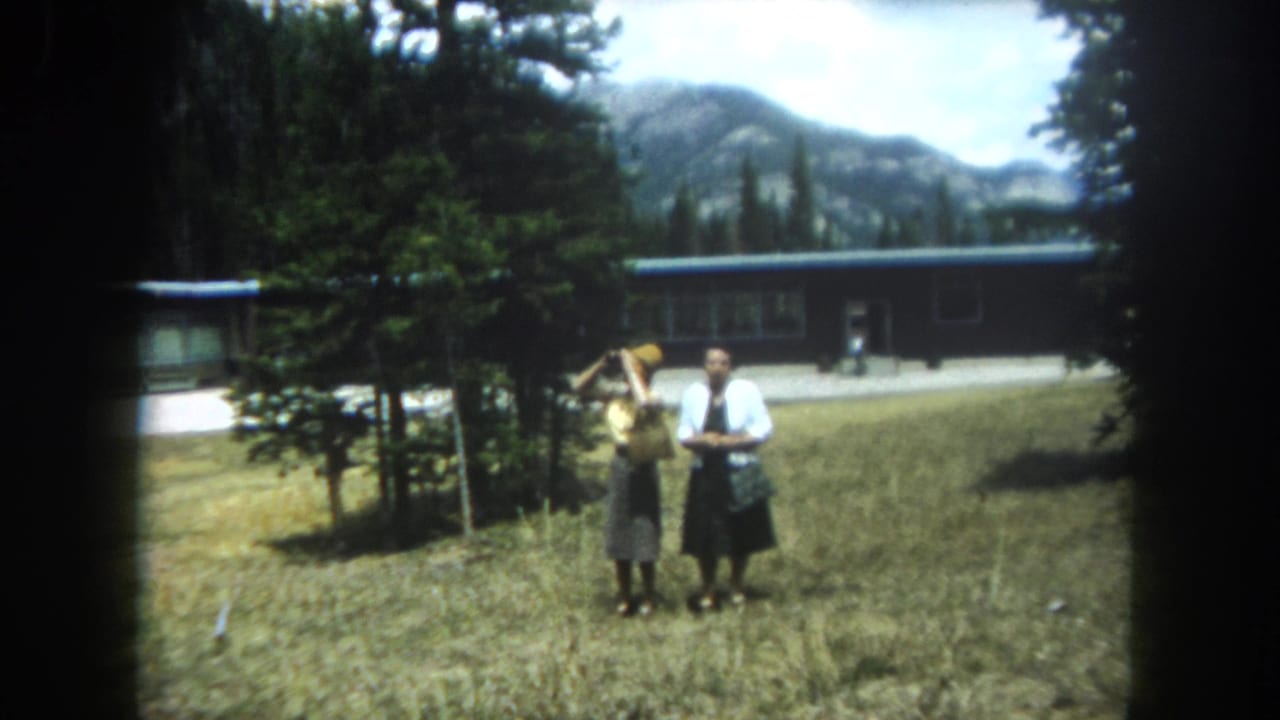 Two women in early 1960s attire in a Summit County, Colorado field — one in yellow top raising what may be a camera, one in white shirt and dark skirt — pine trees and rocky peaks behind.
