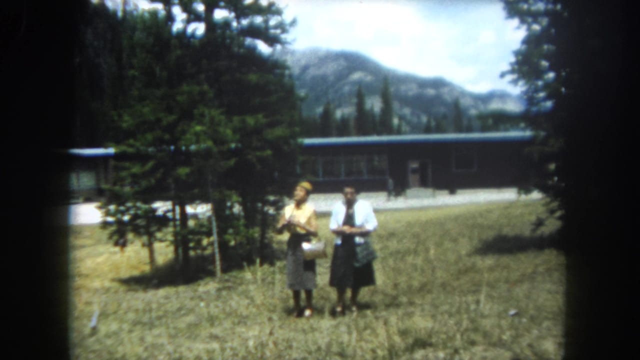 Two women in 1960s summer attire stand together in a Colorado mountain field, a mid-century building with dark roof and rocky mountain peaks visible behind them, Summit County, circa 1962.