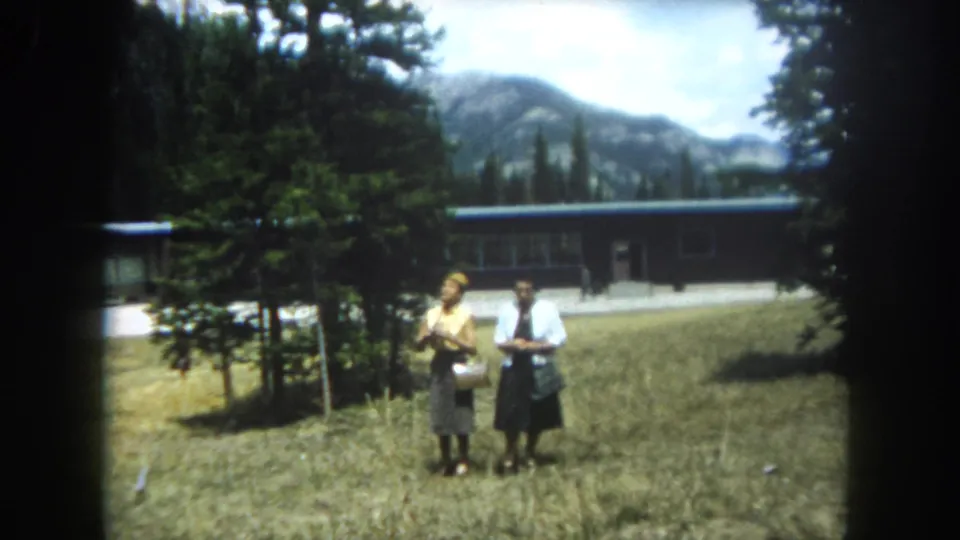 Two women in 1960s summer attire stand together in a Colorado mountain field, a mid-century building with dark roof and rocky mountain peaks visible behind them, Summit County, circa 1962.