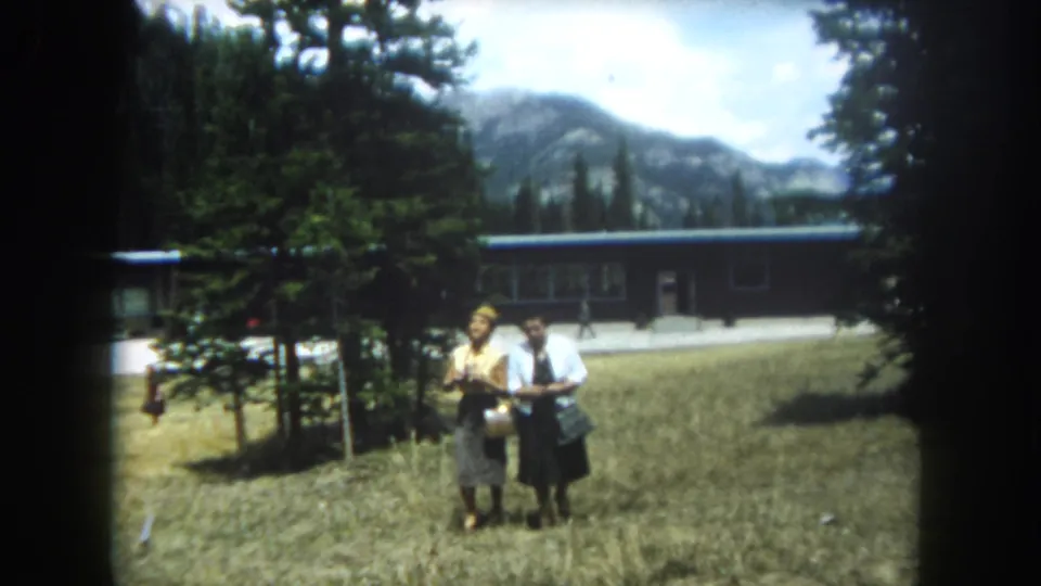 Closer framing of two women — one in yellow, one in white carrying a bag — in a Colorado mountain field, with a lodge-style building and rocky peaks behind, Summit County, circa 1962.