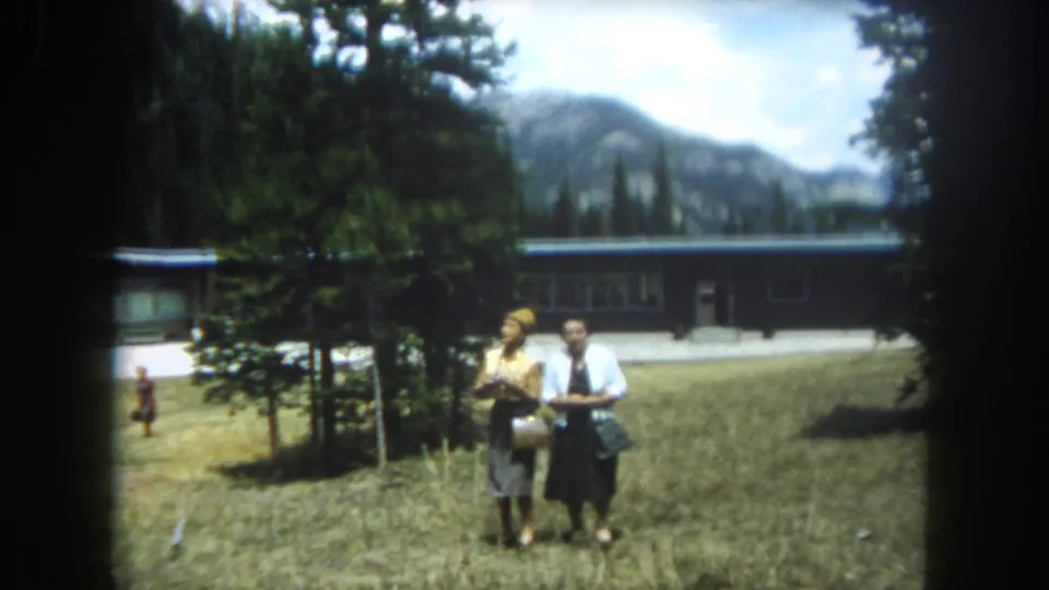 Two women in summer attire stand in a Colorado mountain setting, a mid-century building among conifers and a prominent rocky ridge visible behind them, Summit County, circa 1962.