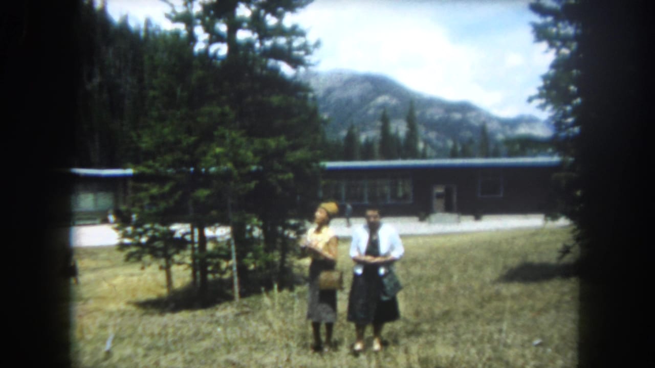 Two women in 1960s dress stand in a sun-lit Rocky Mountain clearing near a flat-roofed building, with conifer forest and peaks behind them, Summit County Colorado circa 1962.
