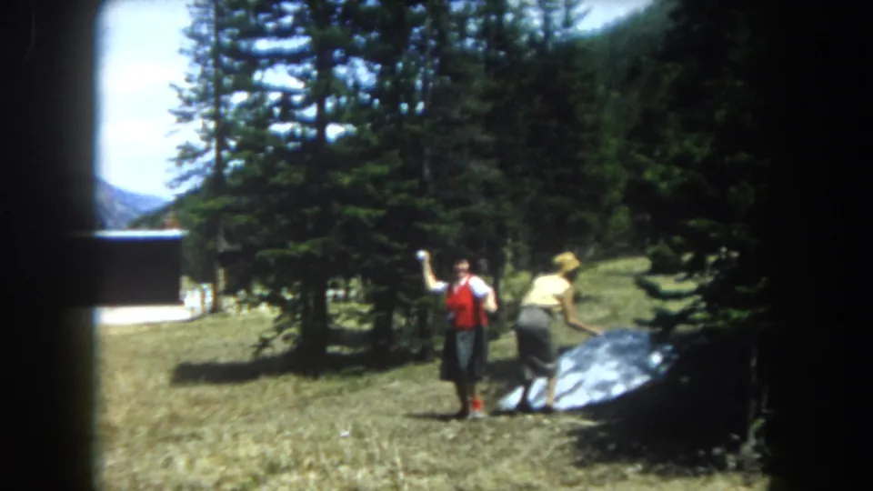 Two women in 1960s folk-influenced dress, one in a red vest over white blouse, handle a large parabolic reflective dish together in a Colorado mountain clearing, circa 1962.