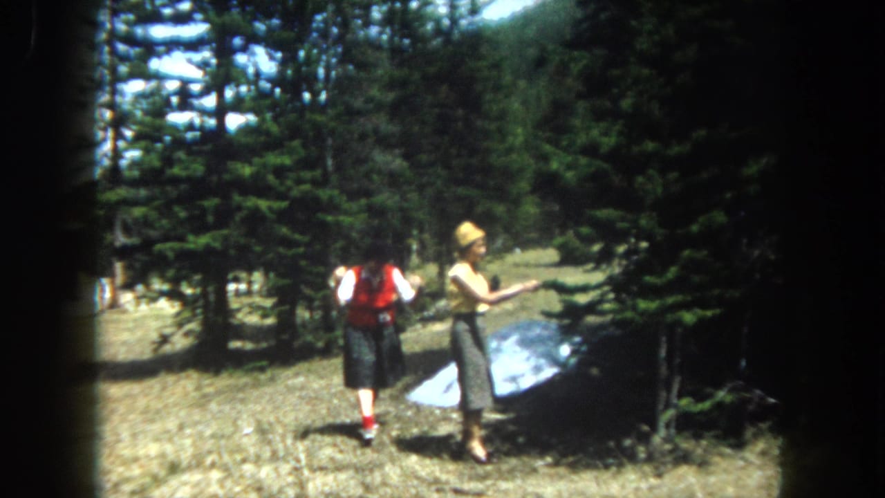 Two women in early-1960s casual dress stand in a rocky high-altitude clearing, likely Summit County Colorado circa 1962, with a flat reflective panel on the ground and snow-dusted pines behind.
