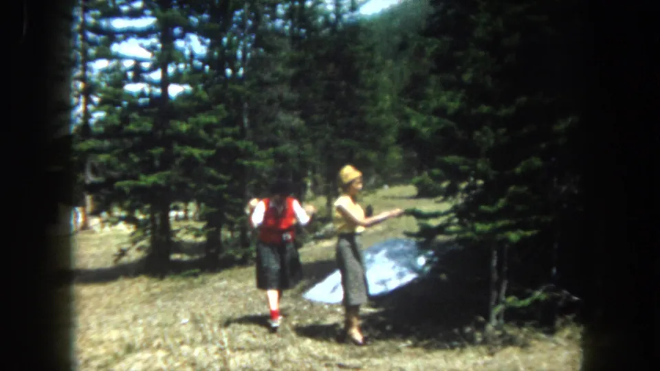Two women in early-1960s casual dress stand in a rocky high-altitude clearing, likely Summit County Colorado circa 1962, with a flat reflective panel on the ground and snow-dusted pines behind.