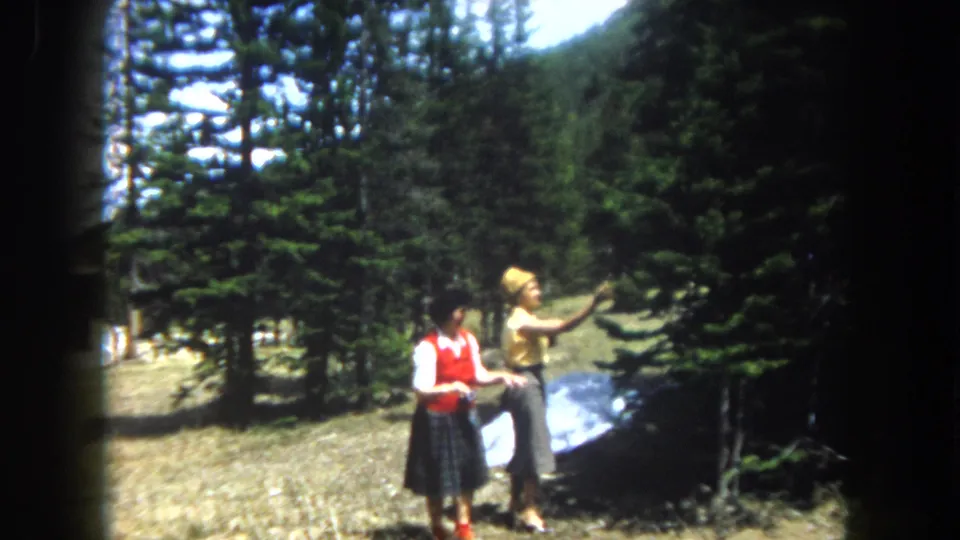 Two women in early-1960s dress stand in a high-altitude clearing in what appears to be Summit County Colorado, with a prominent reflective panel on the ground and snow visible between the pines.