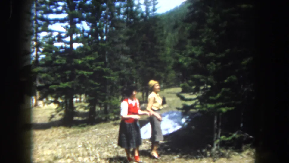 Two women in early-1960s casual attire stand together in a high-elevation pine clearing, likely Summit County Colorado circa 1962, with a large reflective panel on the ground and snow between the trees.