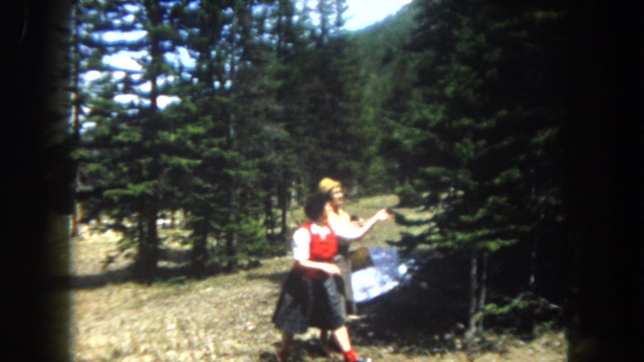 Two women in early-1960s attire, one in a red vest and dark skirt, the other in lighter separates, stand in a pine-forested clearing, likely Summit County, Colorado, c. 1962.