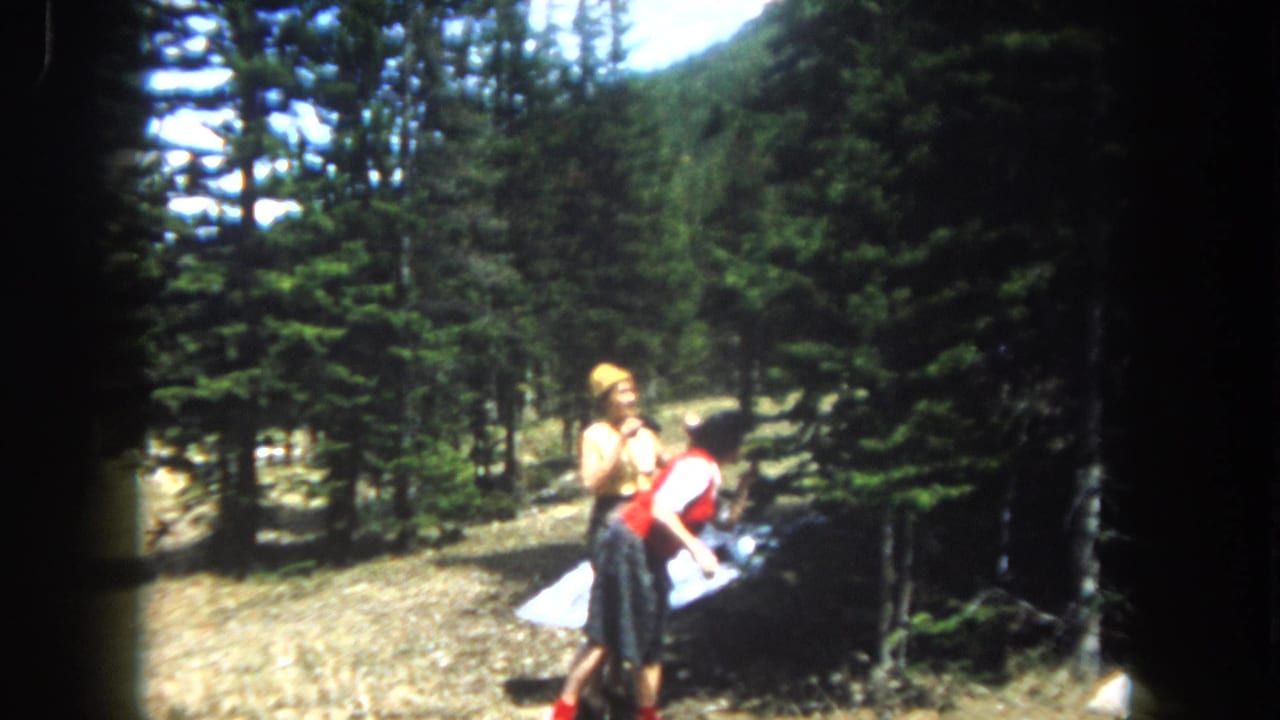 Two women in early-1960s clothing pause in a conifer-framed mountain clearing; one holds what appears to be a folded map or paper, consistent with vacation travel, Summit County, Colorado, c. 1962.