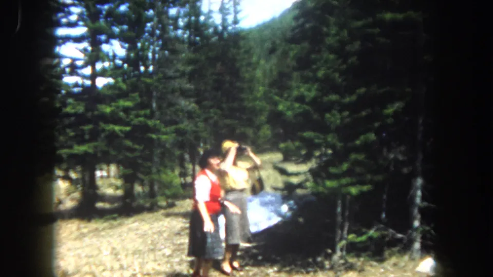 Two women in a pine-forested mountain clearing; one raises an object, possibly binoculars or a camera, to her face, while the other in a red vest looks on, c. 1962 Colorado.