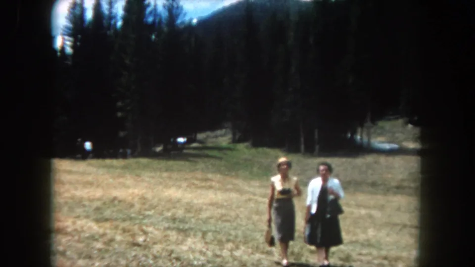 Two women walk toward the camera across an open grassy clearing; a pine forest and mountain ridge are visible behind them, consistent with Summit County, Colorado, c. 1962.