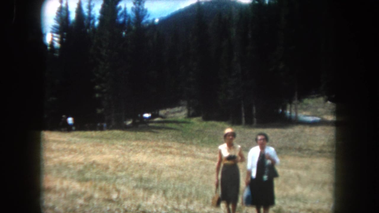 Two adult women pause in a mountain meadow in what appears to be Summit County, Colorado, circa 1962, filmed on 8mm; dense conifers and a ridgeline behind them.