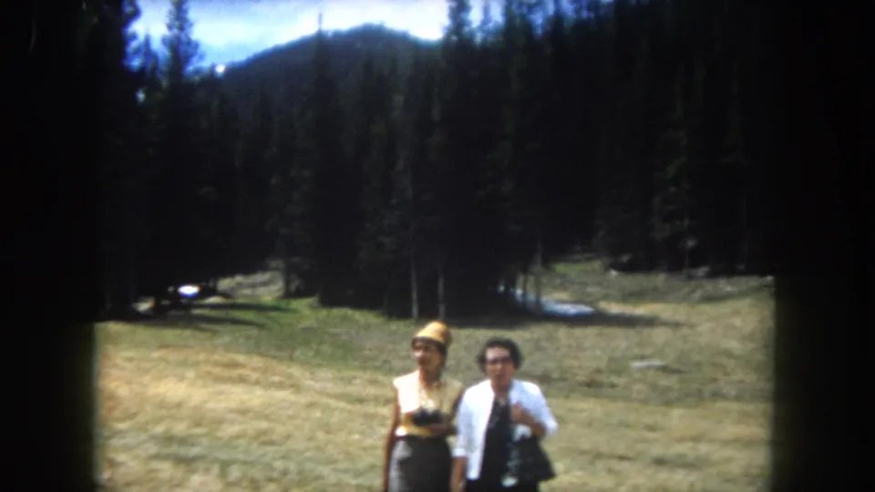 Two adult women approach the camera in an alpine meadow, Summit County, Colorado, circa 1962; dry grass, conifer treeline, and a mountain ridge visible.