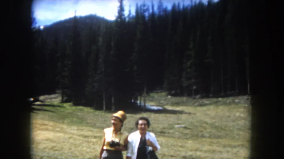 Close framing of two adult women in a Rocky Mountain meadow, circa 1962; one in a yellow straw hat, one in a white cardigan; conifer ridge behind them.