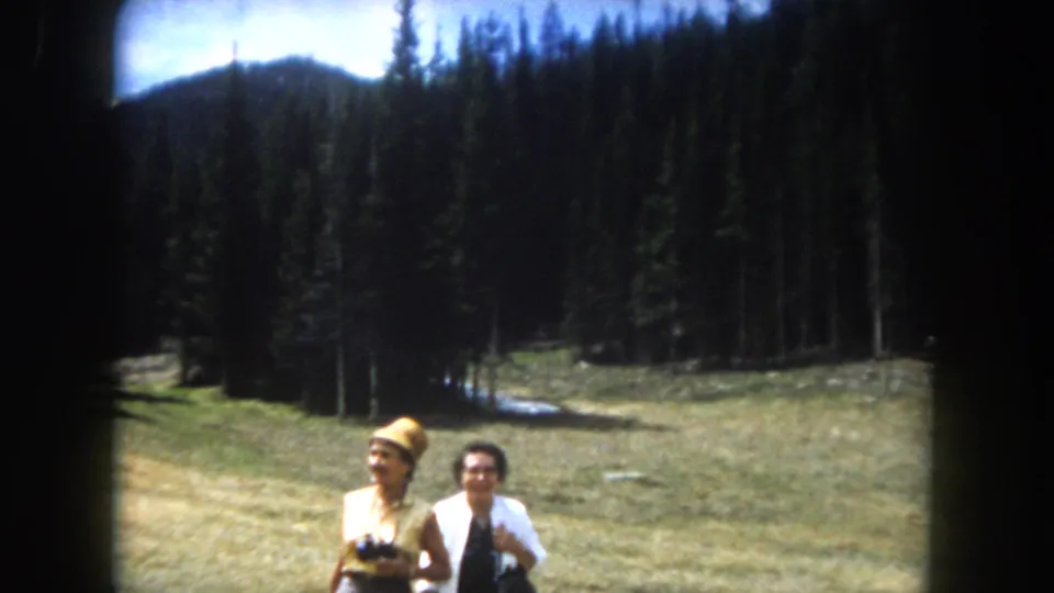 Two women in early-1960s attire pose in an alpine meadow, consistent with Summit County, Colorado, 1962; yellow hat, binoculars, snow-patched peaks visible behind pine forest.