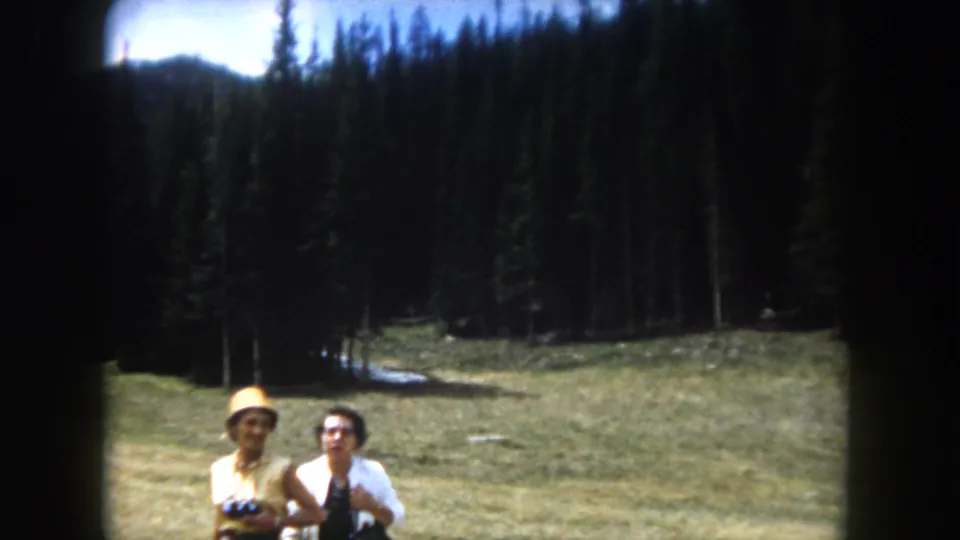 Two women in early-1960s clothing in a mountain meadow, consistent with Summit County, Colorado, 1962; binoculars visible, evergreen treeline and snow-streaked peaks behind.