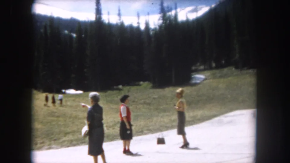 Three women in early-1960s clothing near a paved path in an alpine meadow, consistent with Summit County, Colorado, 1962; snow patches on hillside, pine forest and mountain peaks behind.