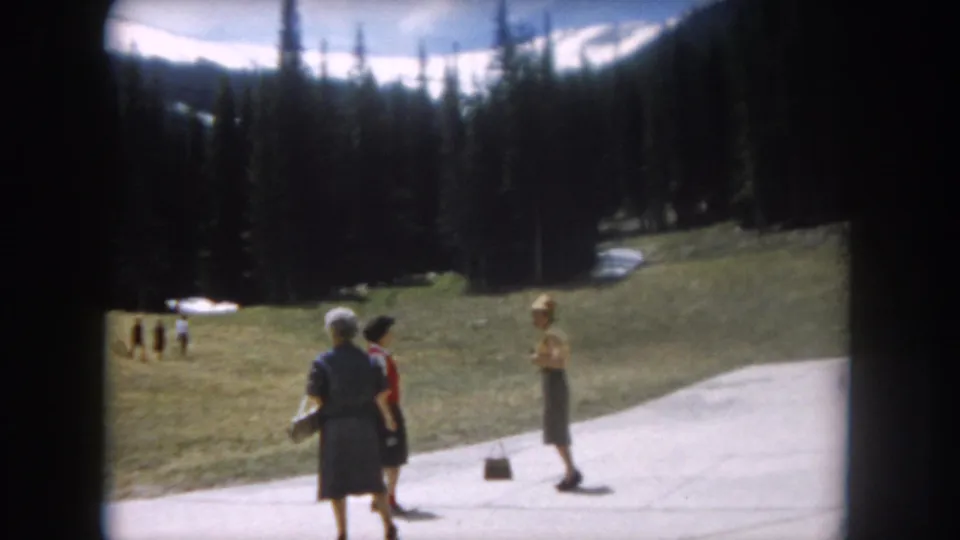 Three women in early-1960s attire near a paved path in an alpine meadow, consistent with Summit County, Colorado, 1962; distant figures visible on slope, snow-patched peaks behind treeline.