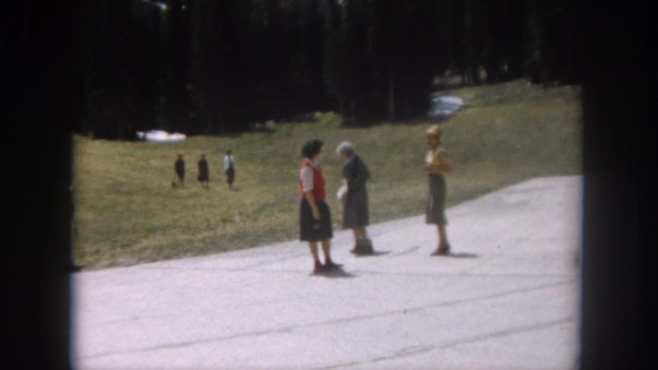 Three women standing on a paved path, conversing; grassy hillside with snow patches and evergreen trees behind them.