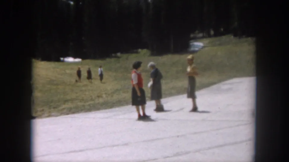 Three women in early-1960s casual dress chat on a paved path at a Colorado mountain recreation area, circa 1962; evergreen trees and snow-dusted hillside in background.