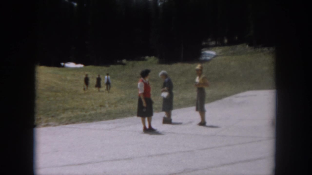 Three women on a paved mountain path in mid-conversation; background walkers visible on grassy hillside amid conifers.