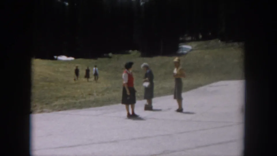 Three women in period dress continue conversing on a paved path, with several figures walking the grassy hillside behind them; Colorado mountain setting, circa 1962.