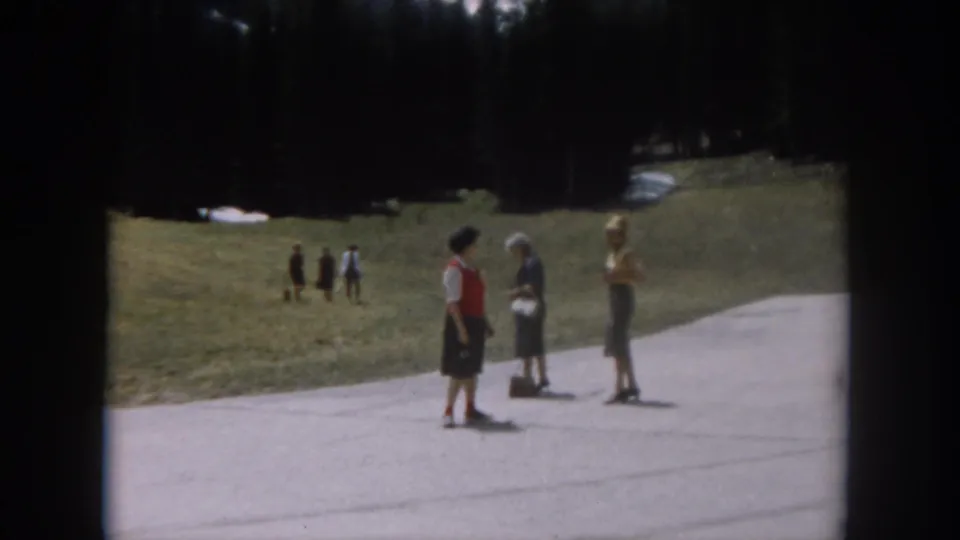 Women in early-1960s attire stand on a paved mountain path, with a snow-patched hillside and dense conifer forest forming the backdrop; Colorado, circa 1962.