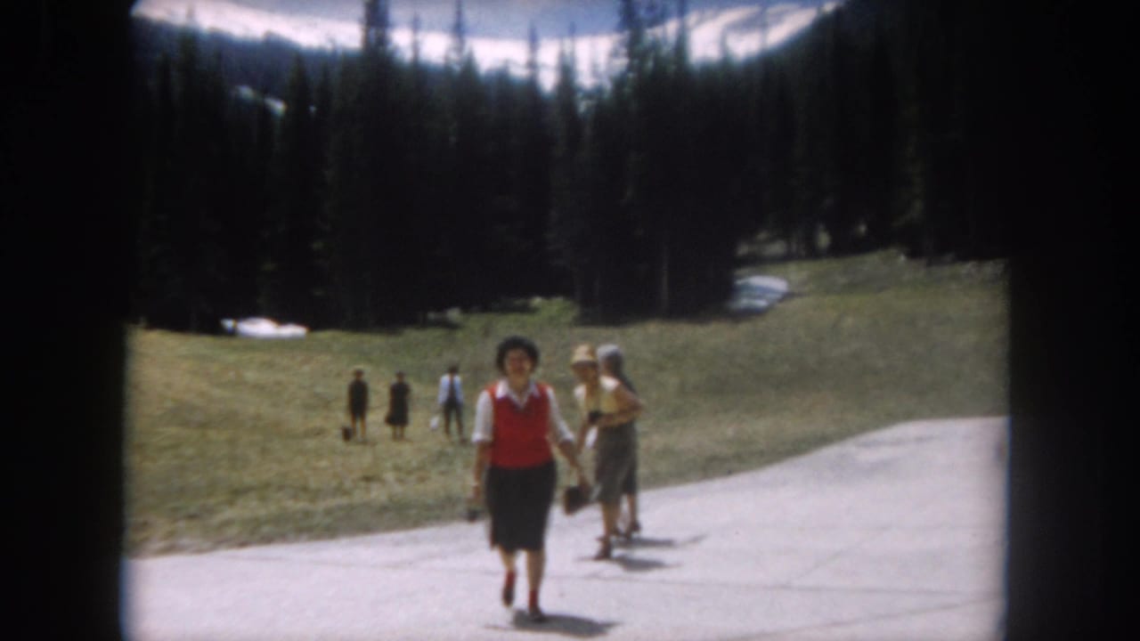 Woman in red vest and dark skirt walks toward camera on paved mountain path; pine forest and snow-patched slopes behind.