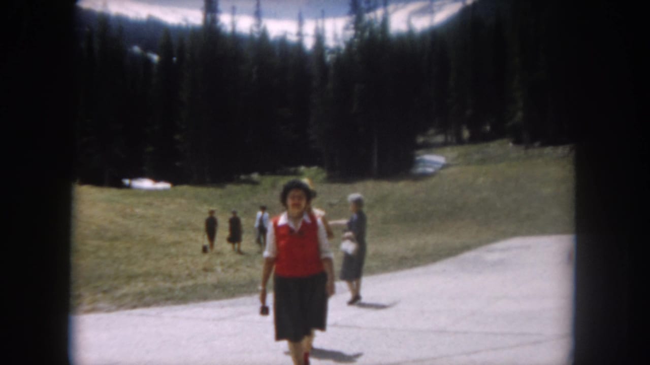 1962 home movie: woman in red vest and dark skirt approaches camera on mountain path in Summit County, Colorado; pine forest and snow patches on slopes visible.