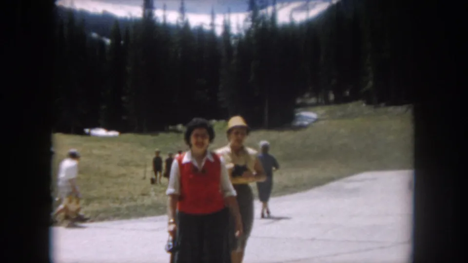 1962 home movie: two women near camera on a mountain path in Summit County, Colorado; pine forest, snow patches, and other visitors visible behind them.