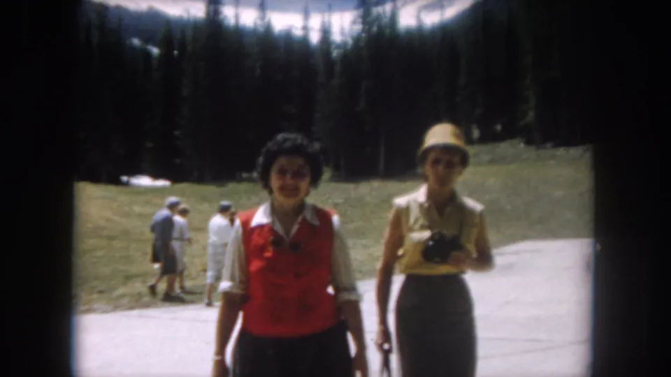 1962 home movie: two women face camera on a mountain path in Summit County, Colorado; woman at right holds a camera; pine forest and snow slopes behind.