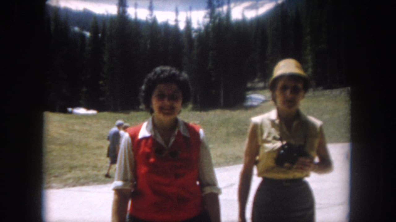 Two women on a paved mountain path — one in a red vest over a white shirt, the other in a straw hat holding a camera — with dense conifer forest behind them.