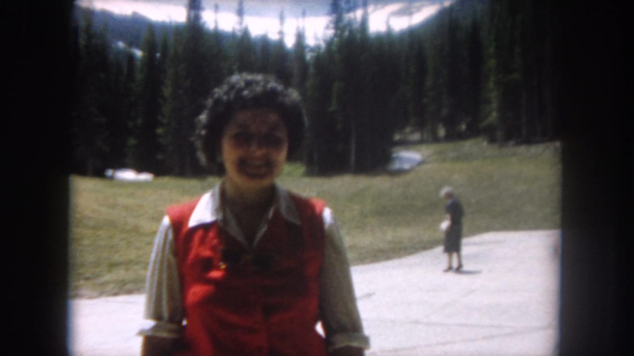 Close shot of a smiling woman in a red vest and white shirt on a mountain path, with evergreen trees, a grassy slope, and a distant visitor visible behind her.