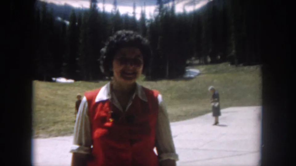 Woman in a bright red vest smiling on a paved resort path at a Colorado mountain location, circa 1962, with other visitors, evergreen trees, and a snow-patched slope behind her.
