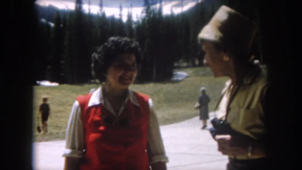 Two women conversing on a Colorado mountain resort path, circa 1962; the companion in a straw hat holds what appears to be a camera, with other visitors and dense conifers visible behind them.