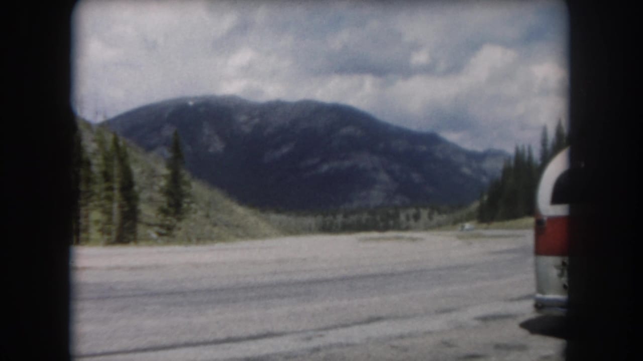 Wide mountain road with red-and-white vehicle ahead, evergreen slopes and rocky peaks under cloudy sky, shot on 8mm film.