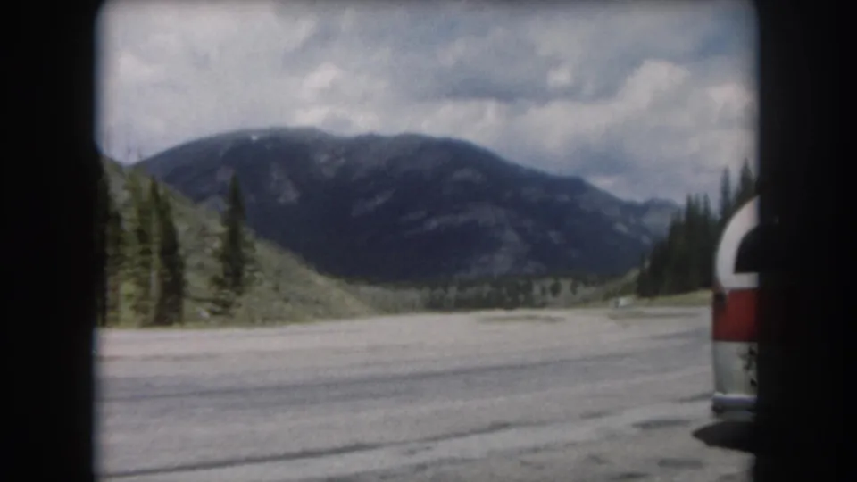 8mm home movie frame circa 1962 showing a mountain highway in Summit County, Colorado, with a red-and-white vehicle and forested peaks.