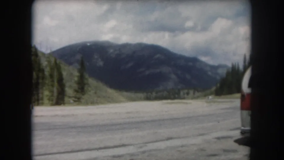 Continuation of 8mm driving sequence circa 1962 showing the same Summit County mountain road and red-and-white vehicle under partial cloud cover.