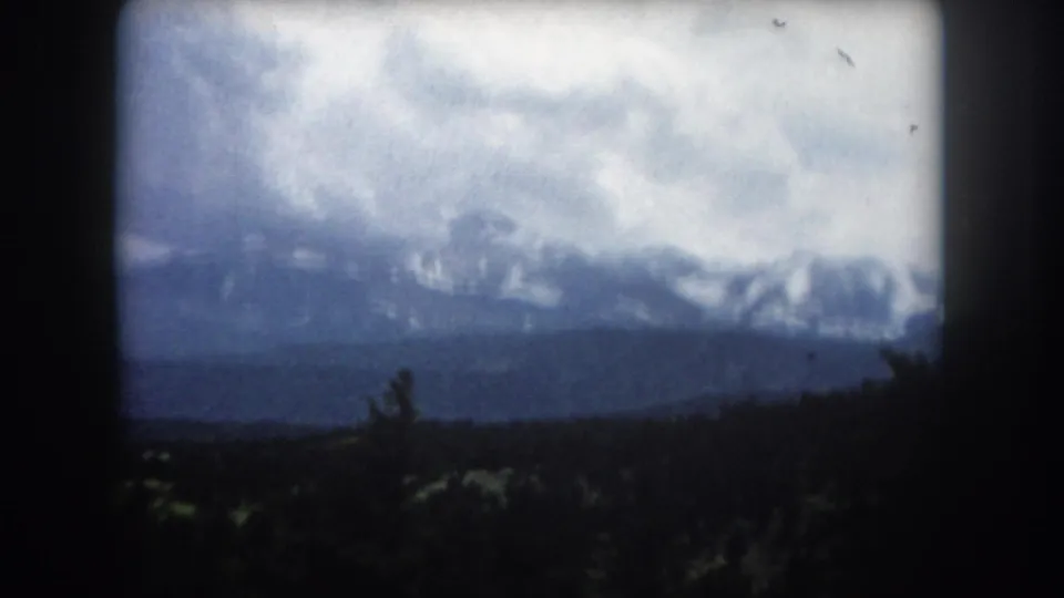 8mm home movie circa 1962 — snow-capped peaks of a Colorado mountain range under dramatic overcast sky, with a conifer silhouette at the frame edge.