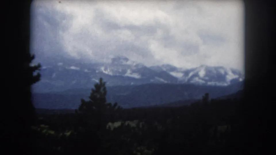 8mm home movie circa 1962 Colorado — wide mountain panorama shot from inside a vehicle, with conifers in the foreground and a dark structural element at frame right.