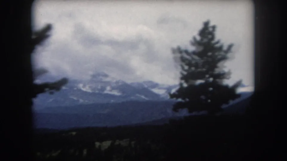 8mm home movie circa 1962 Summit County Colorado — a conifer tree silhouette at right frames snow-capped peaks and an overcast sky.