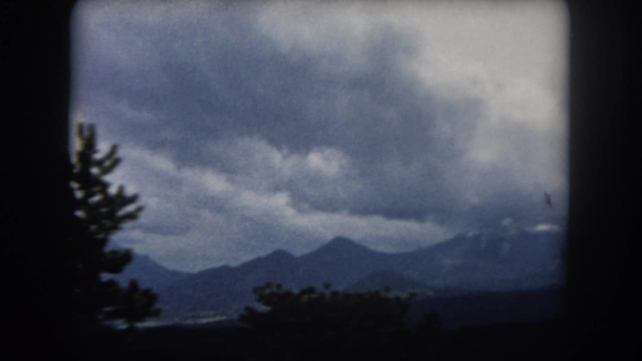 1962 Summit County, Colorado view of layered mountain peaks and a silhouetted evergreen under overcast sky.