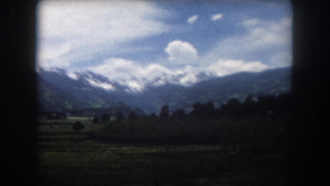 1962 Summit County, Colorado mountain view with distant snowy ridges, a shaded forest foreground, valley floor, clouds, and 8mm frame edges.