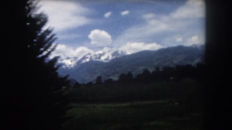 1962 Summit County, Colorado 8mm view with evergreen silhouettes, distant snow-capped mountains, green slopes, and scattered clouds.